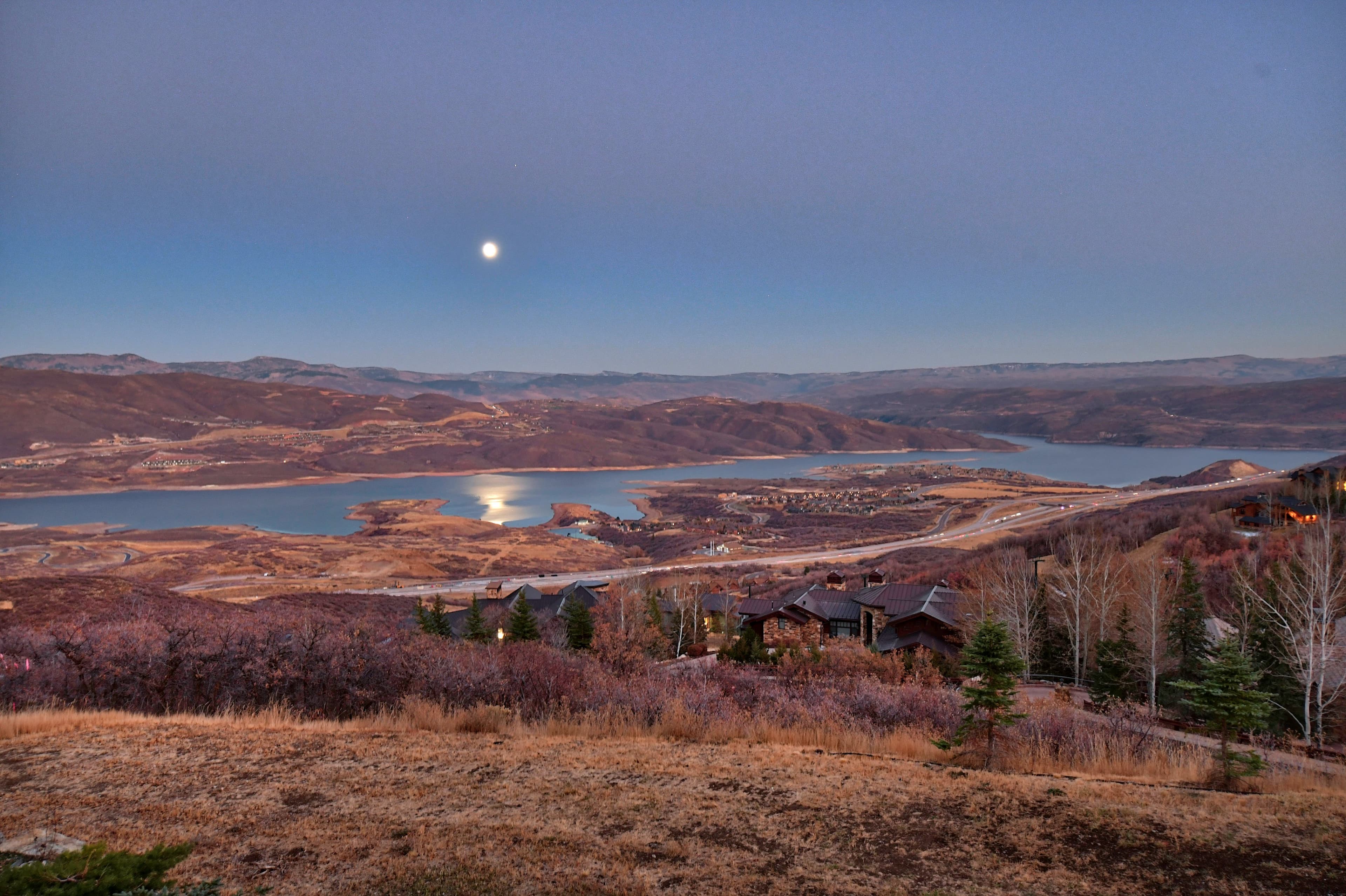 The moon reflects light off the lake at night, with lights from nearby properties visible.